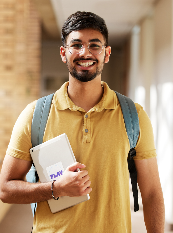 A young man with glasses, wearing a yellow polo shirt and backpack, smiles while holding a tablet labeled "PLAY" in a hallway.
